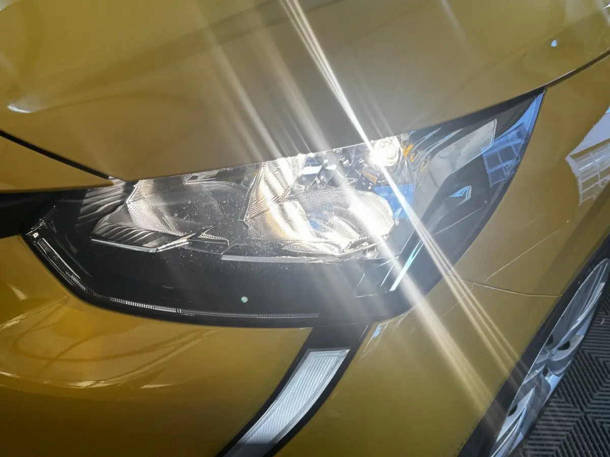 Gros plan sur le phare avant droit d’une Peugeot 208 jaune 2023 avec reflets lumineux sur la carrosserie.