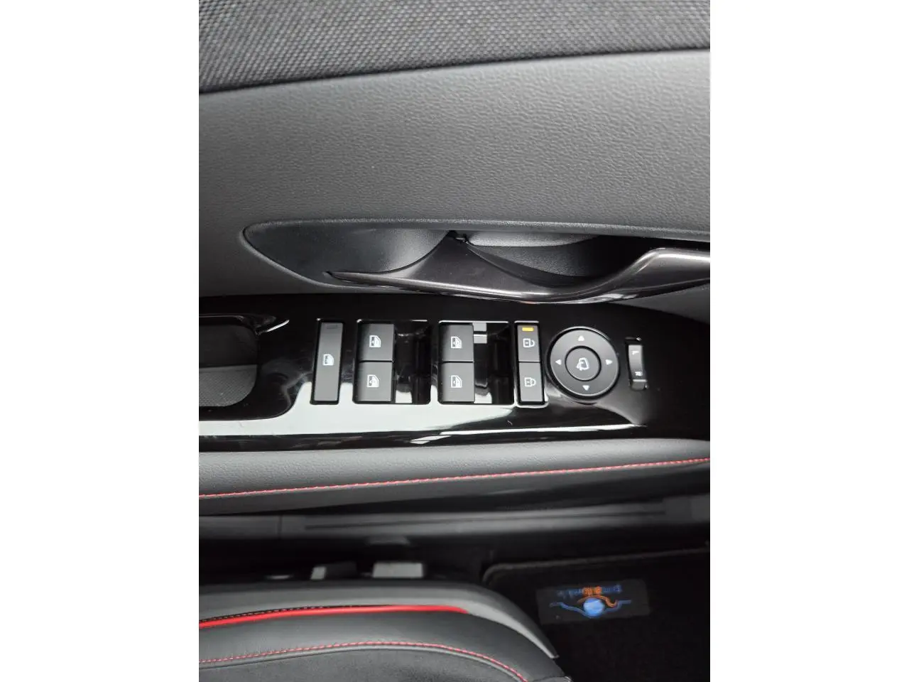 Gros plan sur la commande des vitres et rétroviseurs noirs brillants avec surpiqûres rouges sur la porte côté gauche du Hyundai Tucson 2025.