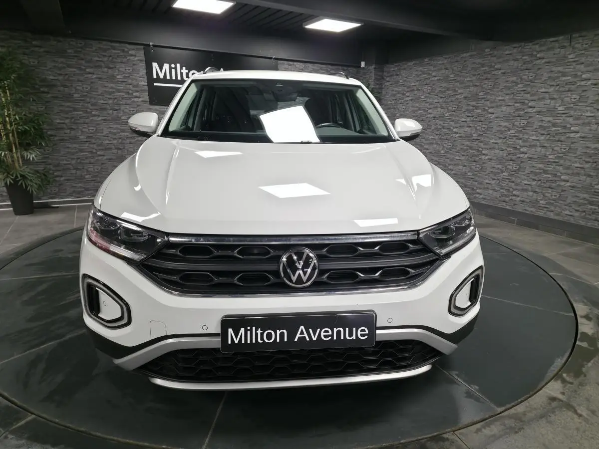Vue de face d'un Volkswagen T-Roc blanc avec calandre noire et plaque "Milton Avenue" en intérieur showroom.