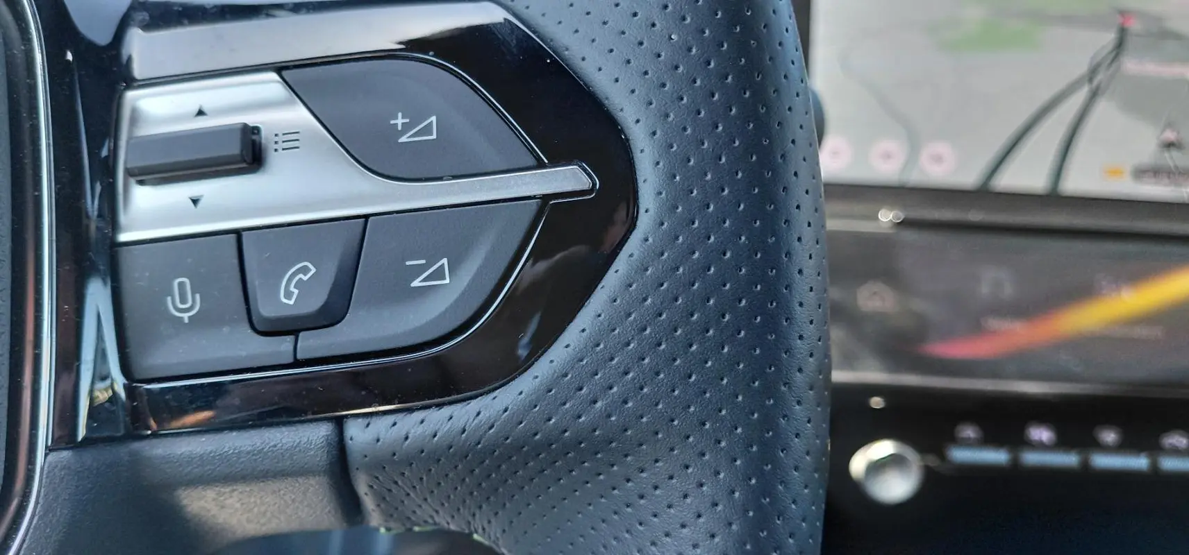 Gros plan sur les commandes gauche du volant cuir perforé d'une Peugeot 308 Hybrid 145 e-DCS6 GT gris, avec cockpit numérique en arrière-plan.