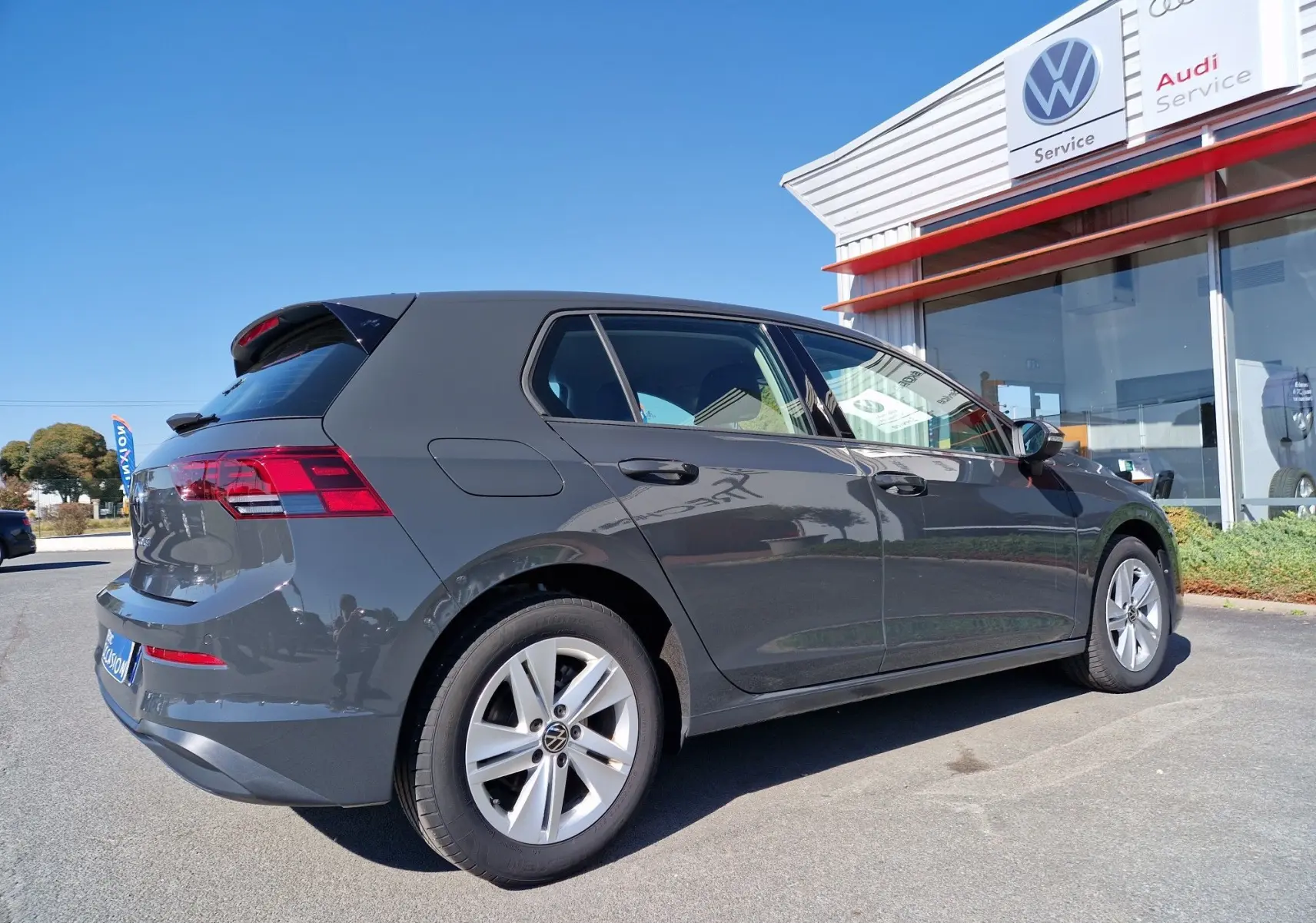 Volkswagen Golf A8 gris Urano vue 3/4 arrière côté gauche, garée devant un service Volkswagen et Audi.