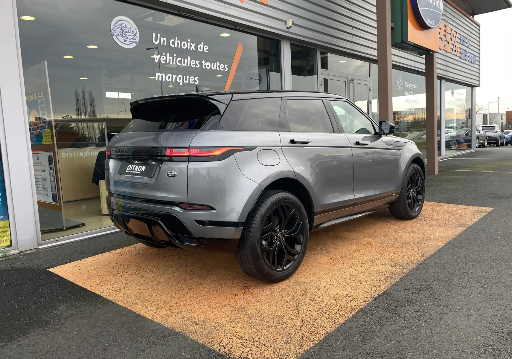 Range Rover Evoque 200D SE R-Dynamic gris Eiger en 3/4 arrière, roues noires et toit noir, stationné devant un showroom.