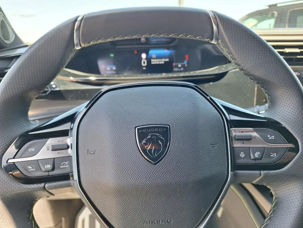 Vue rapprochée du volant cuir noir de la Peugeot 308 Hybrid 145 e-DCS6 GT avec commandes intégrées et tableau de bord numérique flou.