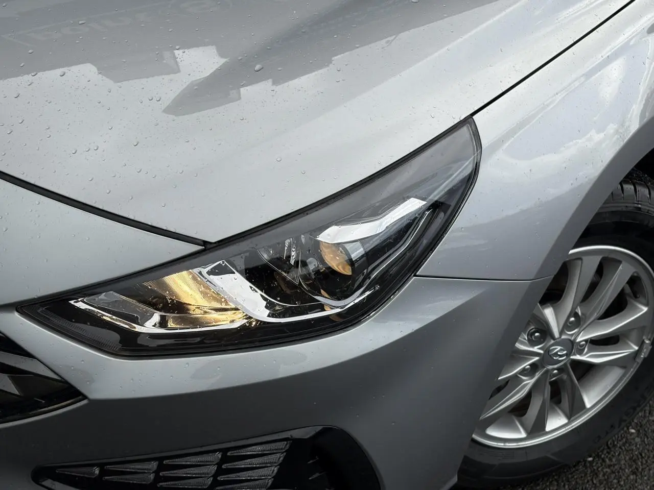 Gros plan sur le phare avant droit allumé et la jante alliage d’une Hyundai i30 SW T-GDI 120 Modern gris argent sous la pluie.