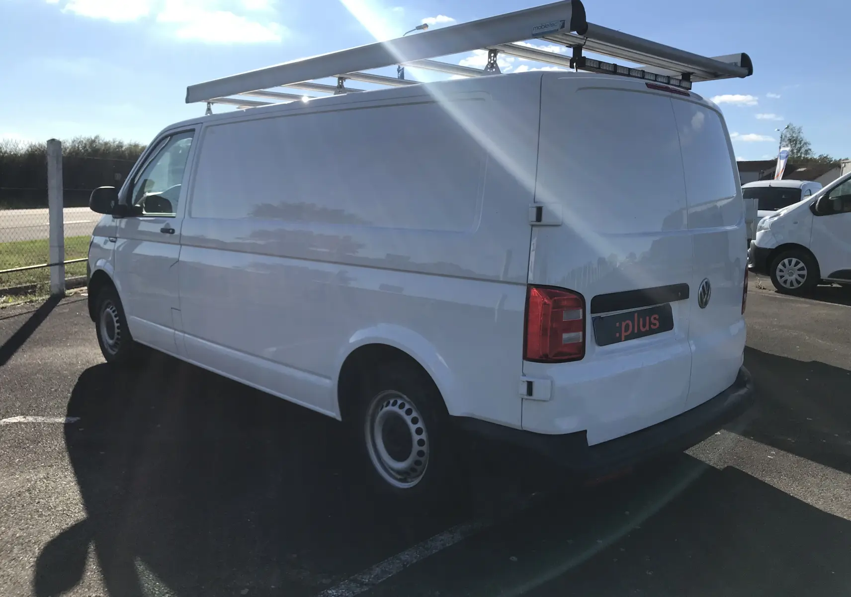 Vue 3/4 arrière droite d'un Volkswagen Transporter blanc équipé d'un porte-échelle sur le toit, stationné en extérieur.