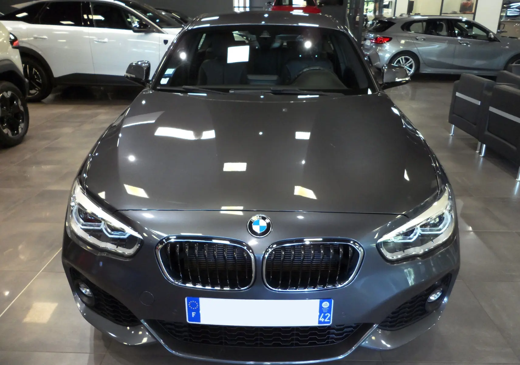 Vue frontale d'une BMW Série 1 F21 LCI gris métallisé en showroom, mettant en valeur sa calandre double et ses phares LED.