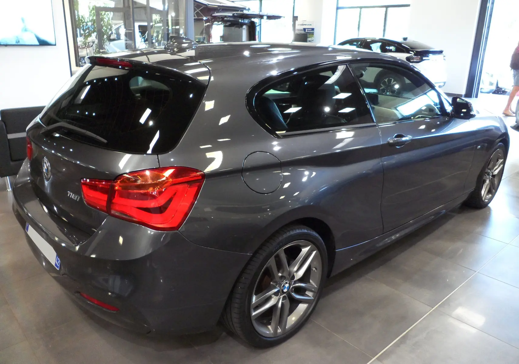 BMW Série 1 F21 LCI 118i gris vue 3/4 arrière droit en showroom avec jantes alliage et feux LED rouges.