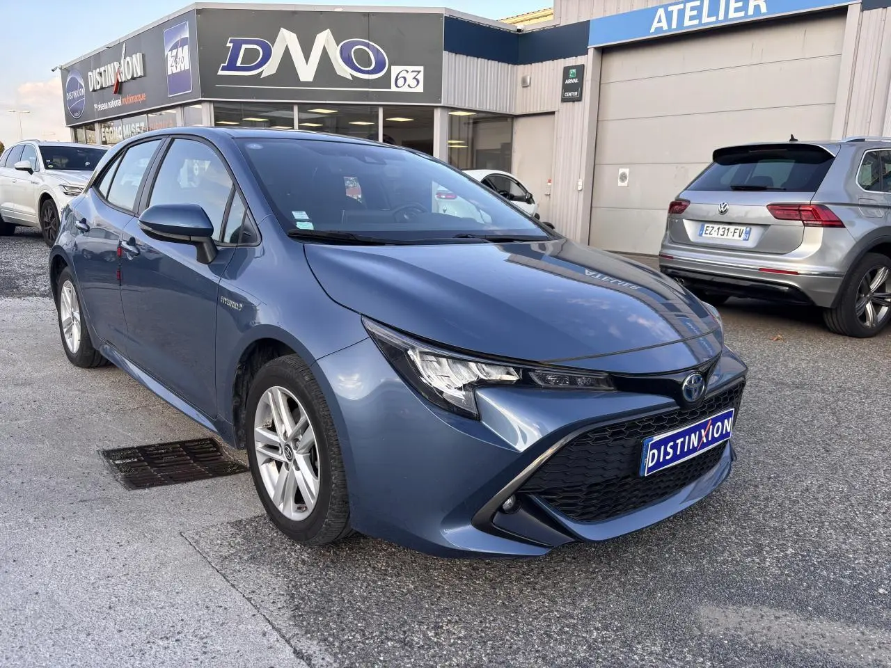 Toyota Corolla hybride bleu clair vue 3/4 avant droit, calandre noire et phares effilés, stationnée devant un atelier.