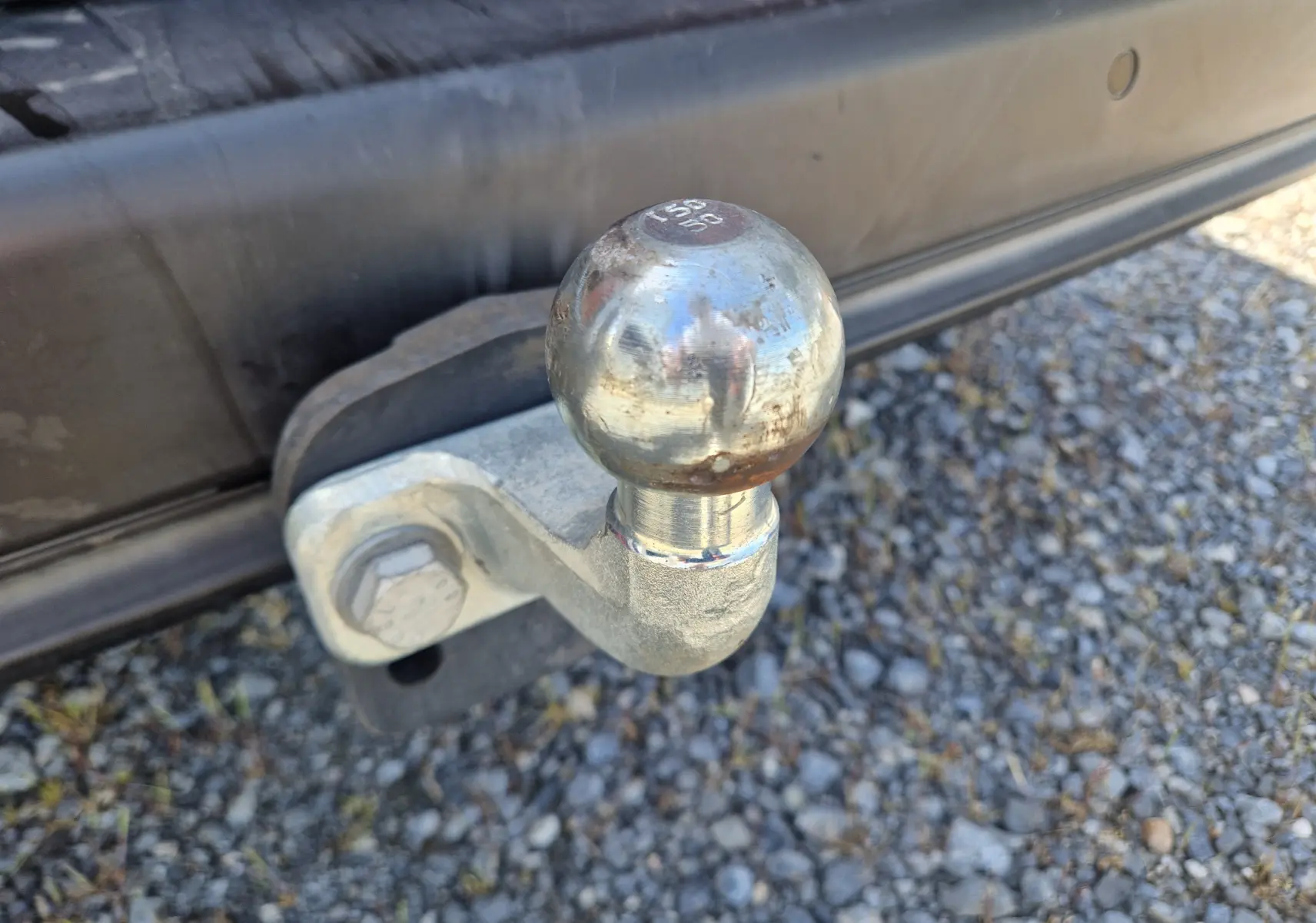 Gros plan sur la boule d’attelage métallique fixée à l’arrière d’un Citroën Berlingo blanc, sur un sol gravillonné.