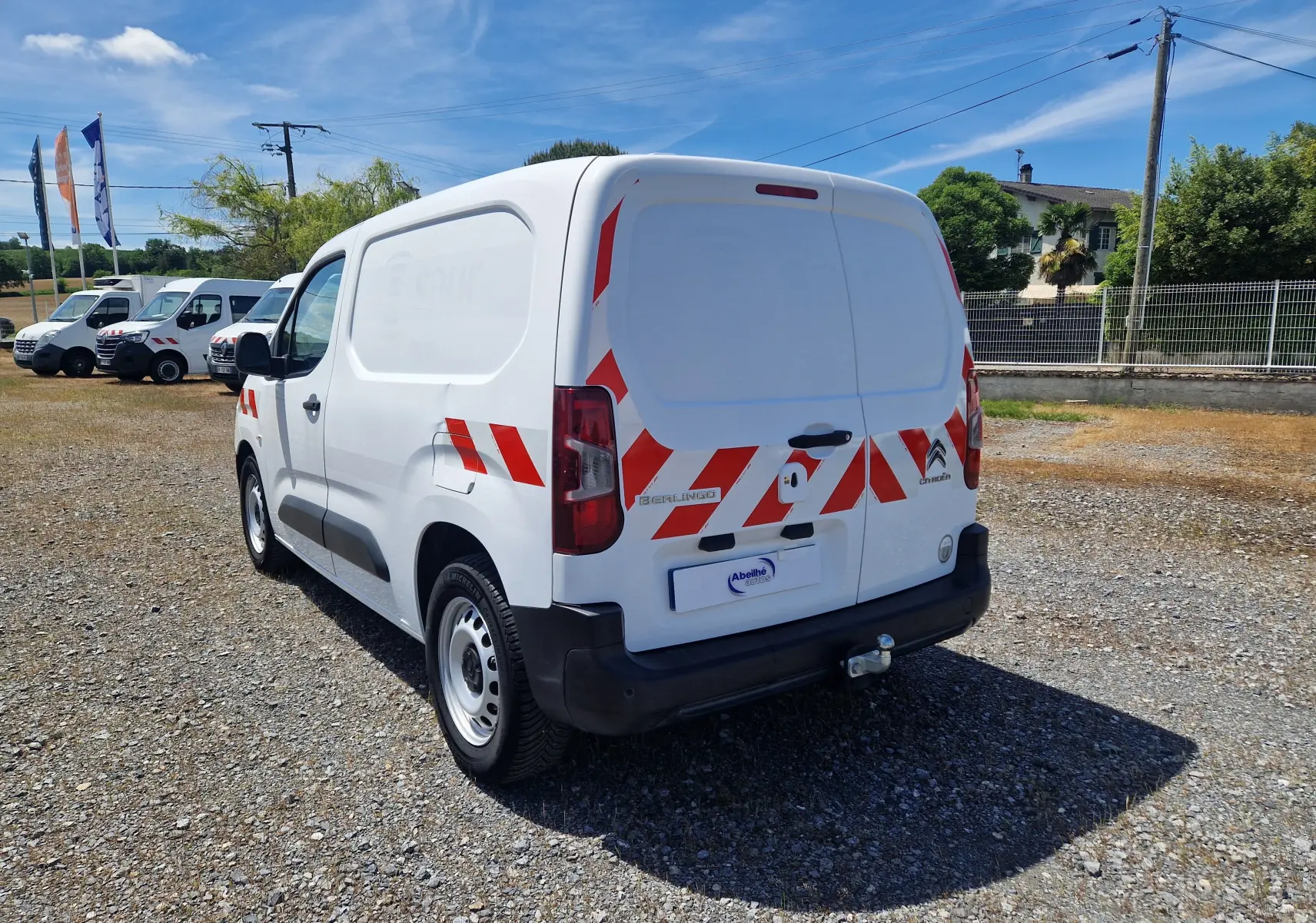 Vue 3/4 arrière droite d’un utilitaire Citroën Berlingo blanc avec bandes rouges réfléchissantes et attelage.