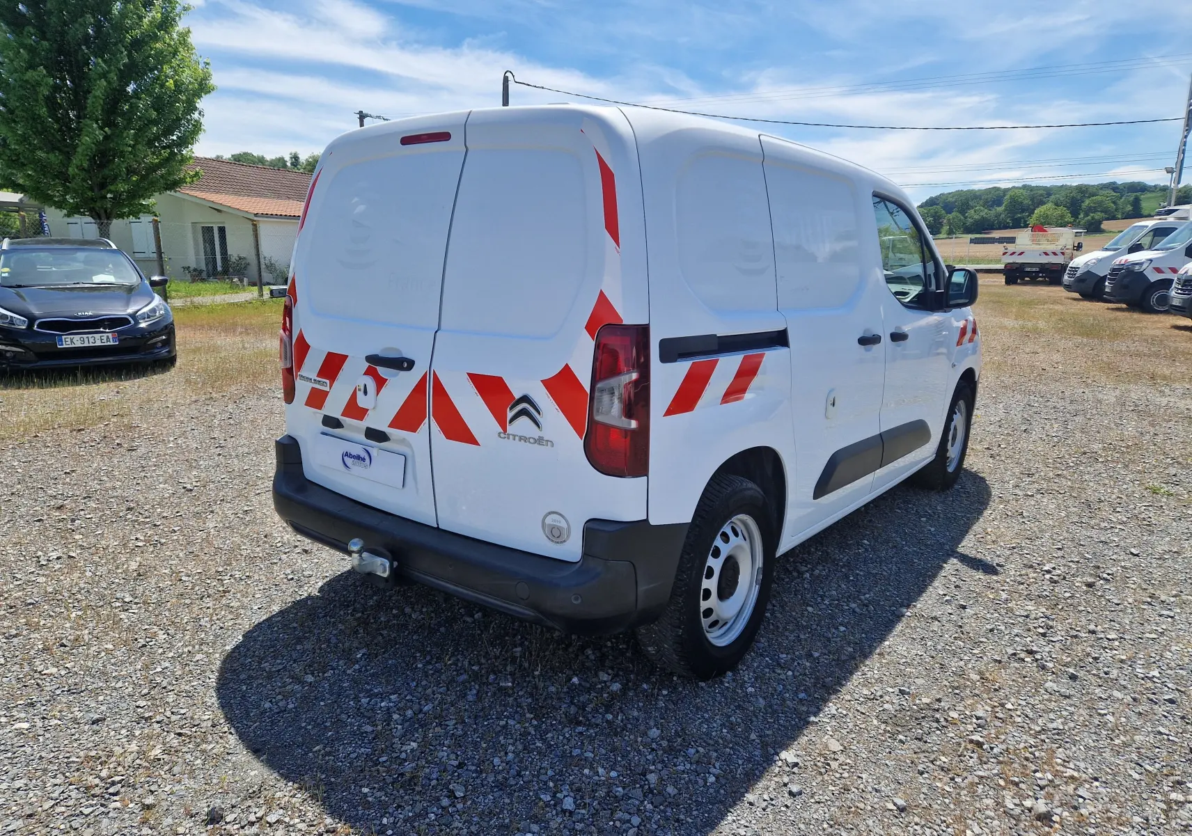 Vue 3/4 arrière droite d’un Citroën Berlingo blanc utilitaire avec bandes réfléchissantes rouges et gyrophare sur le toit.