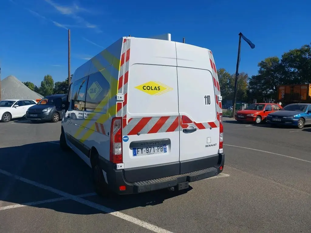 Vue 3/4 arrière droite du Renault Master blanc avec marquages rouges et jaunes Colas, gyrophare et plaque française.