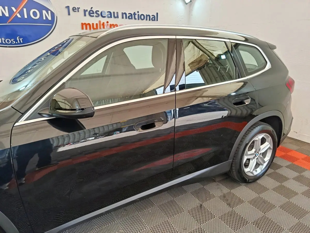 Vue latérale droite d’un BMW X1 noir 2023 en intérieur, avec rétroviseur rabattable et jantes visibles.