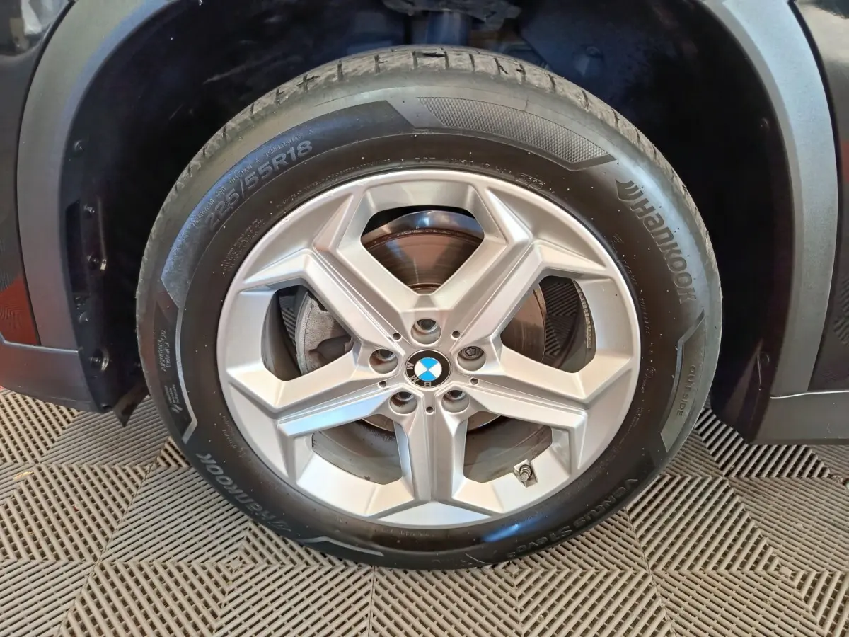 Gros plan sur la roue avant droite avec jante argentée et logo BMW, partie noire du BMW X1 visible en arrière-plan.
