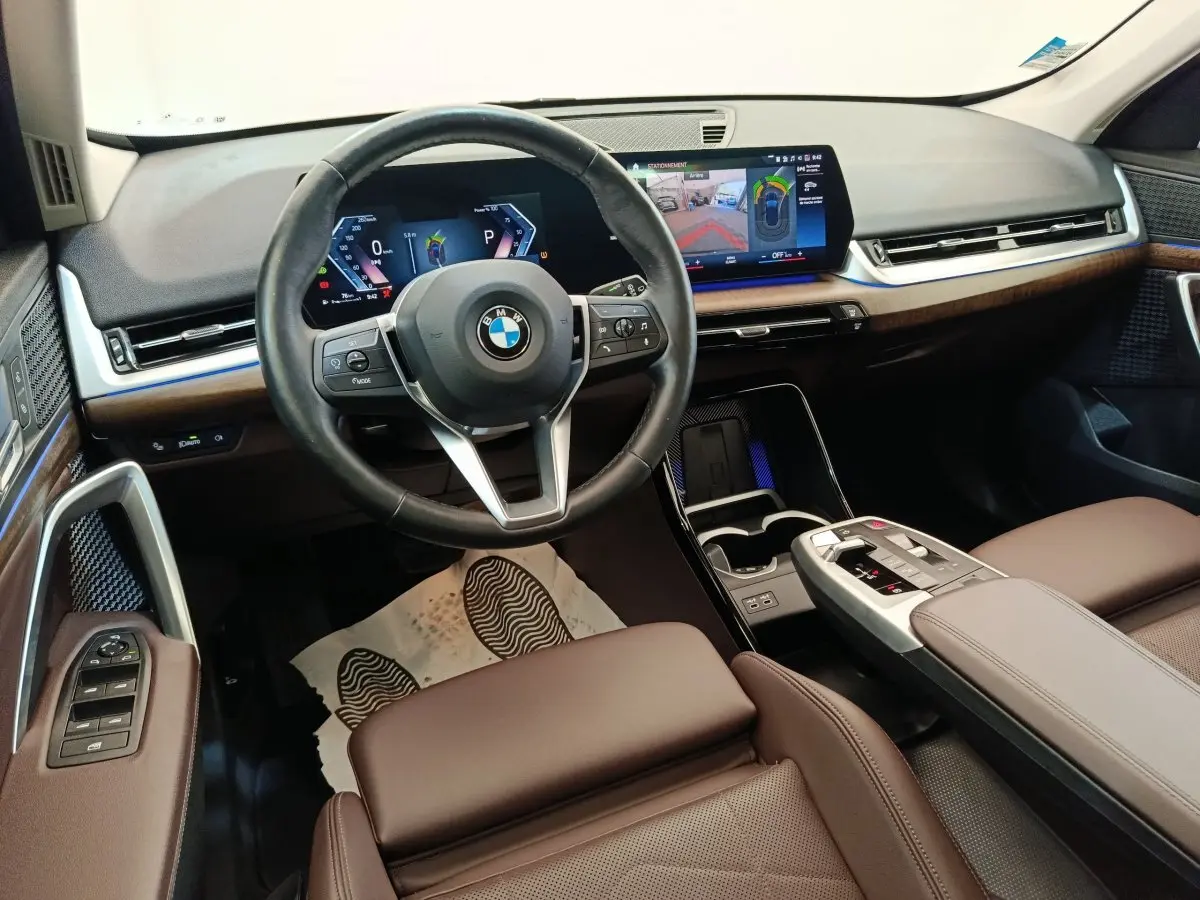Vue intérieure avant du BMW X1 2023 avec volant cuir, écran tactile large et finitions bois marron.