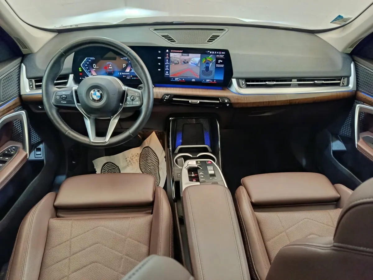 Vue intérieure avant du BMW X1 2023 avec sièges cuir marron, tableau de bord moderne et écran tactile large.