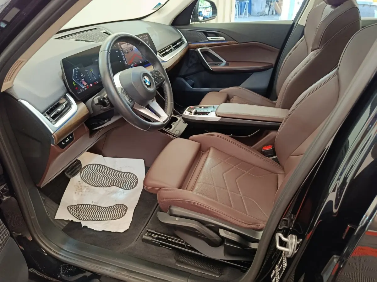 Vue intérieure côté conducteur de la BMW X1 noire 2023, sièges cuir marron et tableau de bord avec écran tactile.