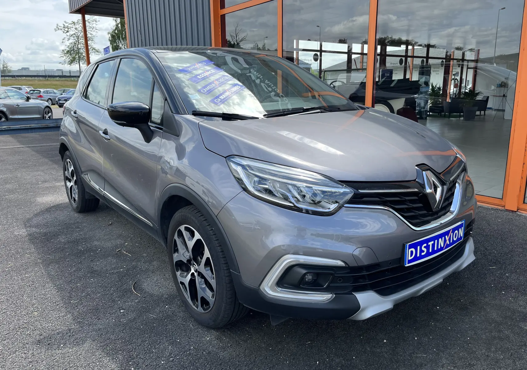 Renault Captur gris en 3/4 avant droit, avec calandre noire et jantes alliage distinctives, stationné devant un bâtiment vitré.