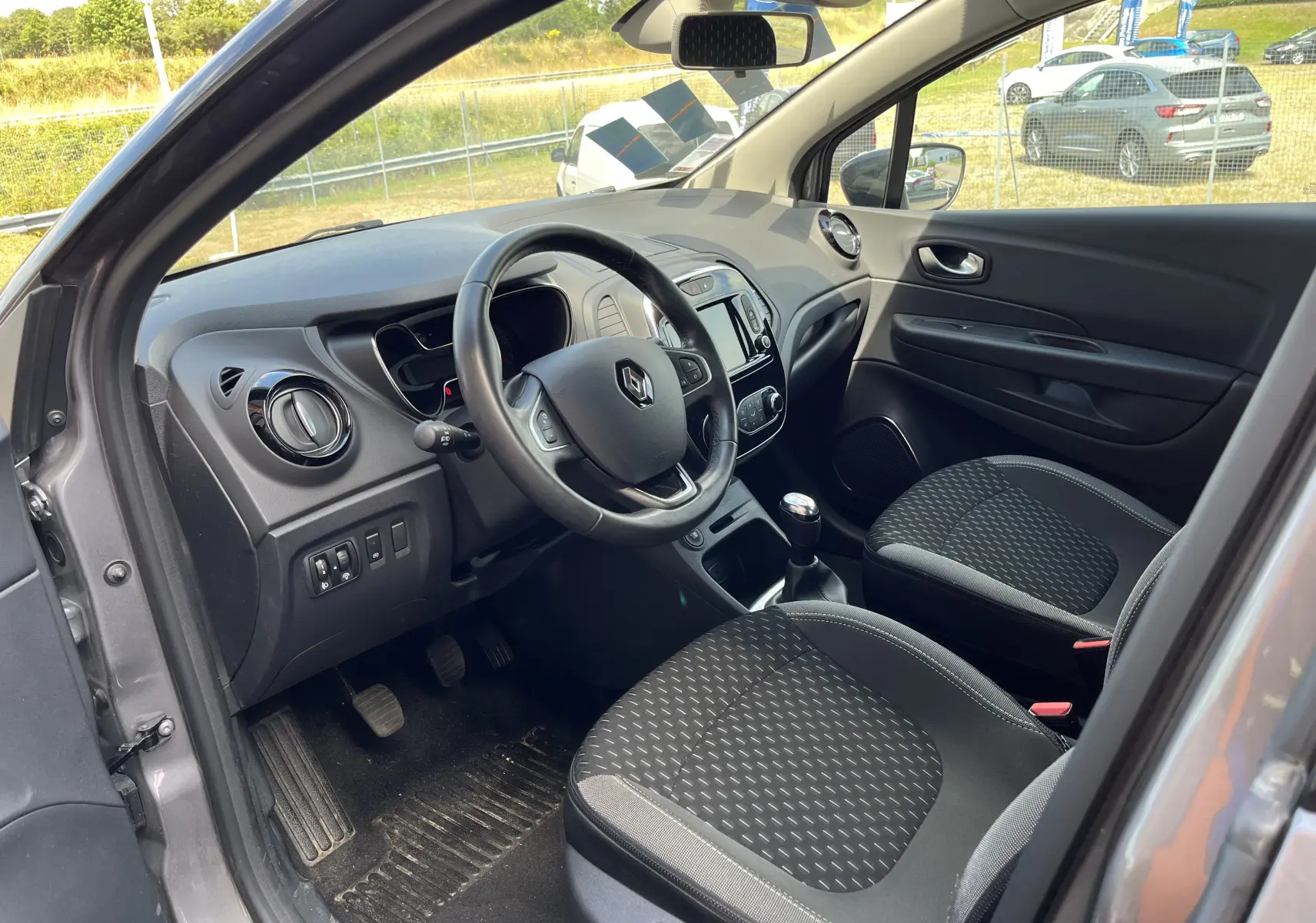 Intérieur avant gauche du Renault Captur gris 2019, volant cuir, tableau de bord avec écran tactile et sièges tissu noirs.