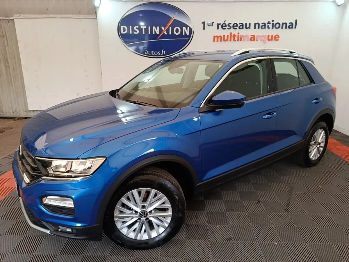 Volkswagen T-Roc bleu en 3/4 avant droit, avec jantes argentées et barres de toit blanches visibles en intérieur showroom.