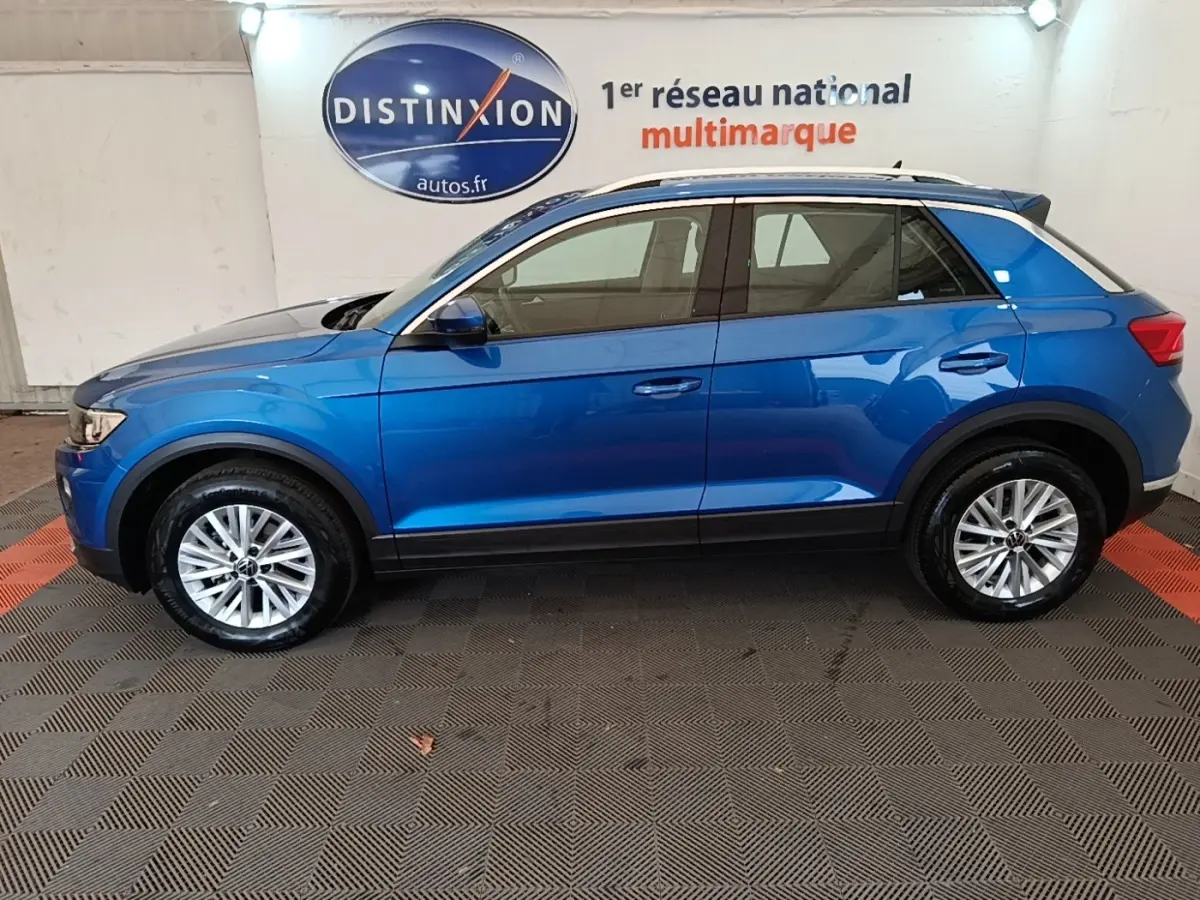 Vue latérale droite du Volkswagen T-Roc bleu 2020, avec jantes alliage et barres de toit noires visibles en intérieur showroom.