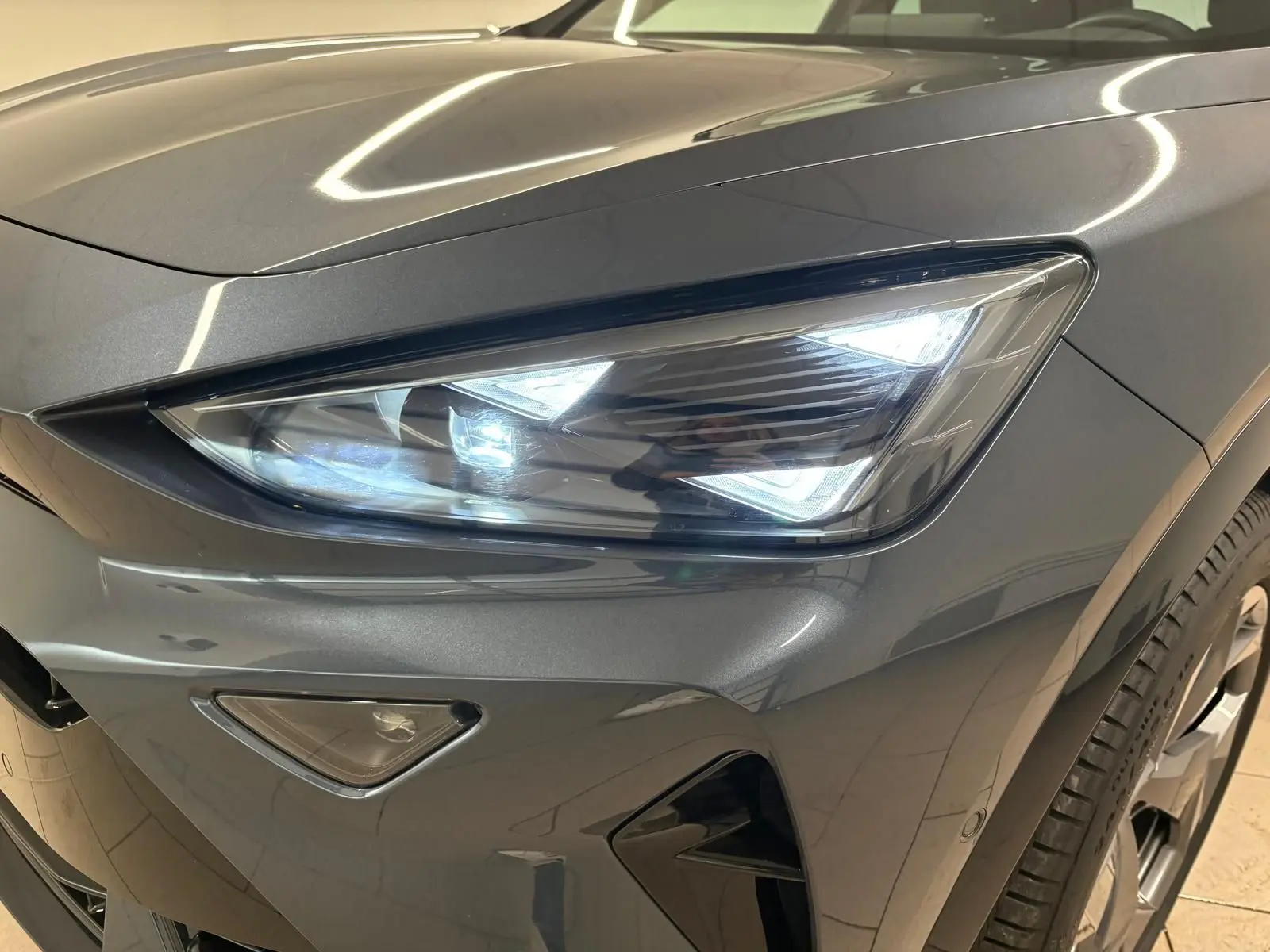 Gros plan sur le phare avant droit allumé d'un CUPRA Formentor gris magnétique avec détails du pare-chocs et roue.