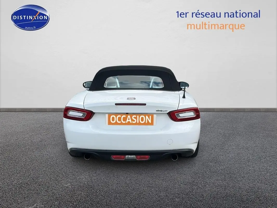 Vue arrière d'une FIAT 124 Spider blanche avec capote noire, feux arrière allongés et double sortie d'échappement.