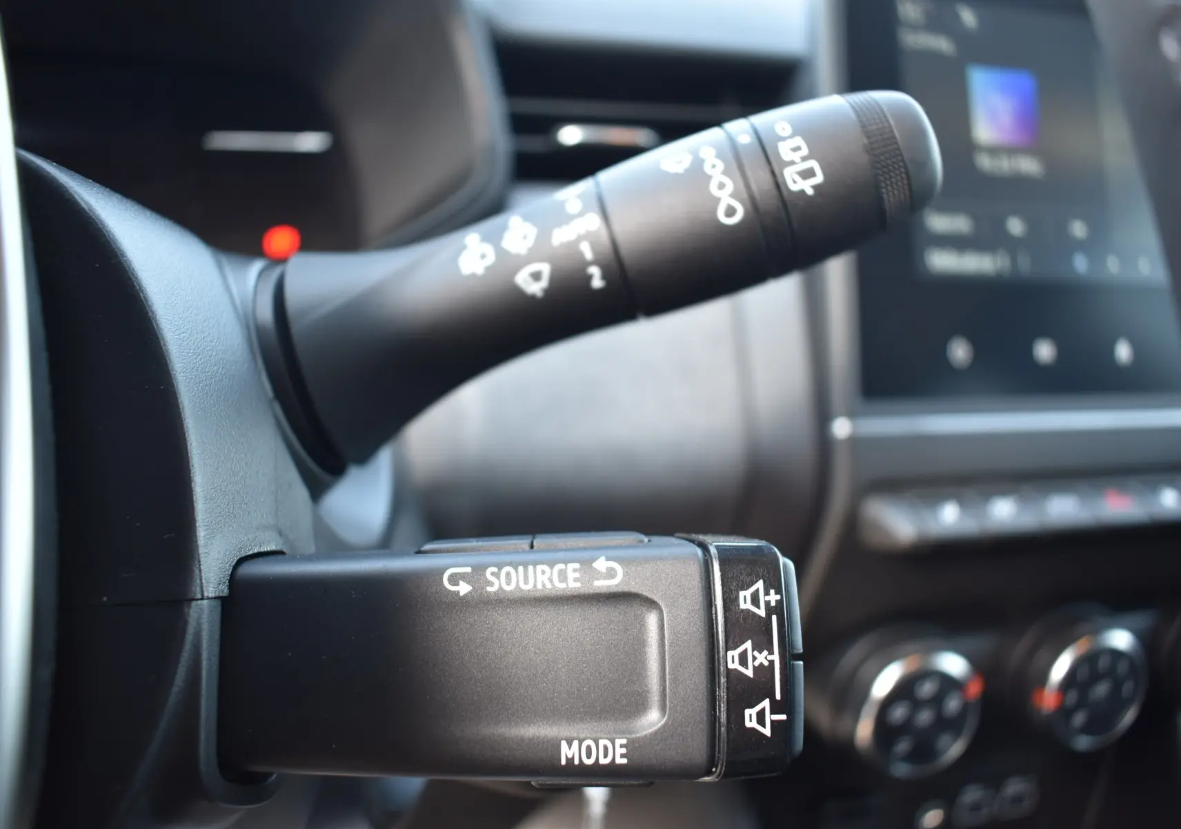 Gros plan sur la commande multimédia au volant de la Renault Clio V gris, avec tableau de bord et commandes clim visibles en arrière-plan.