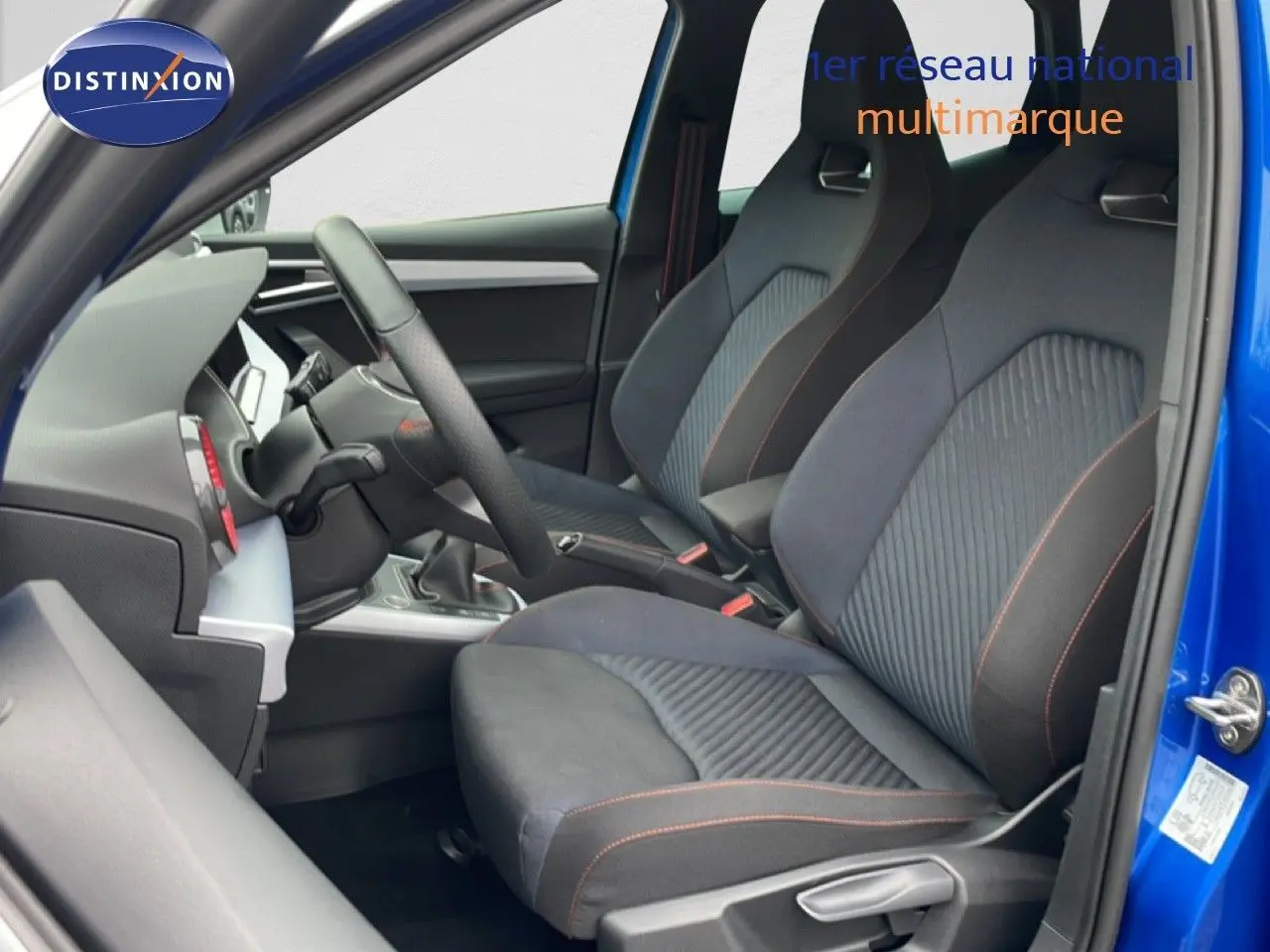 Vue intérieure côté conducteur du SEAT Arona bleu saphir avec sièges tissu noir et surpiqûres orange, volant cuir multifonction.
