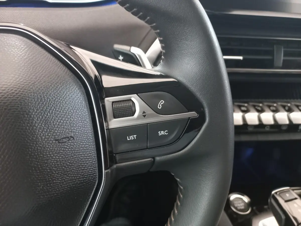 Gros plan sur les commandes au volant cuir noir du Peugeot 3008 2022, avec tableau de bord et touches piano visibles en arrière-plan.