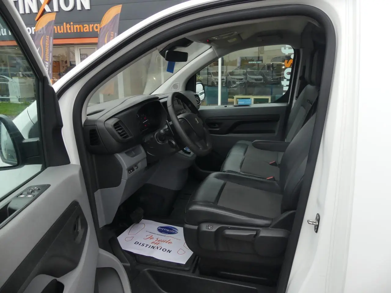 Vue intérieure côté conducteur du Peugeot Expert Fourgon blanc, montrant les sièges avant en tissu et tableau de bord noir.