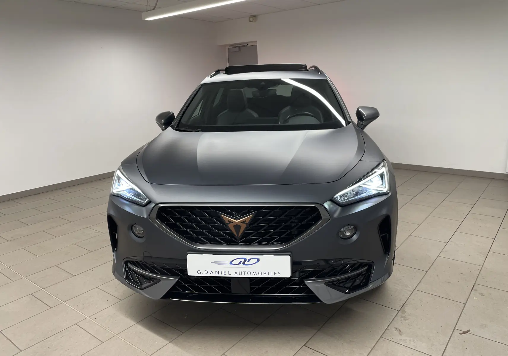 Vue avant d'un CUPRA Formentor 1.5 TSI 150 DSG7 VZ-LINE gris magnétique mat avec phares allumés en intérieur.