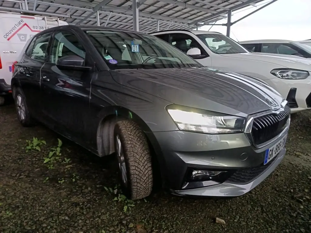 Vue 3/4 avant gauche d'une Skoda Fabia gris graphite 2022 avec phares allumés, stationnée sur sol gravillonné.
