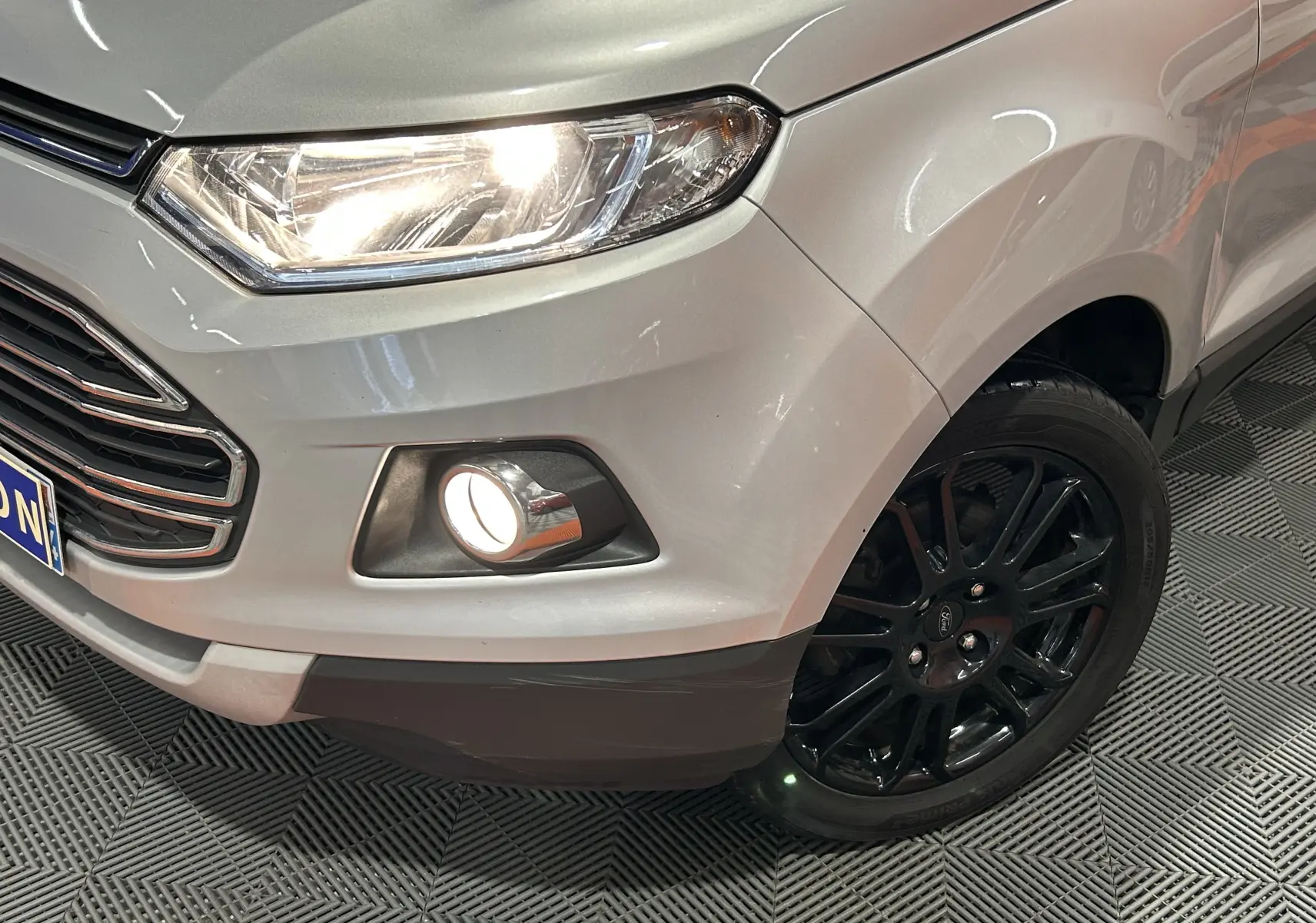 Gros plan sur l'avant droit gris du Ford EcoSport 2018 avec phare et antibrouillard allumés, jante noire visible.