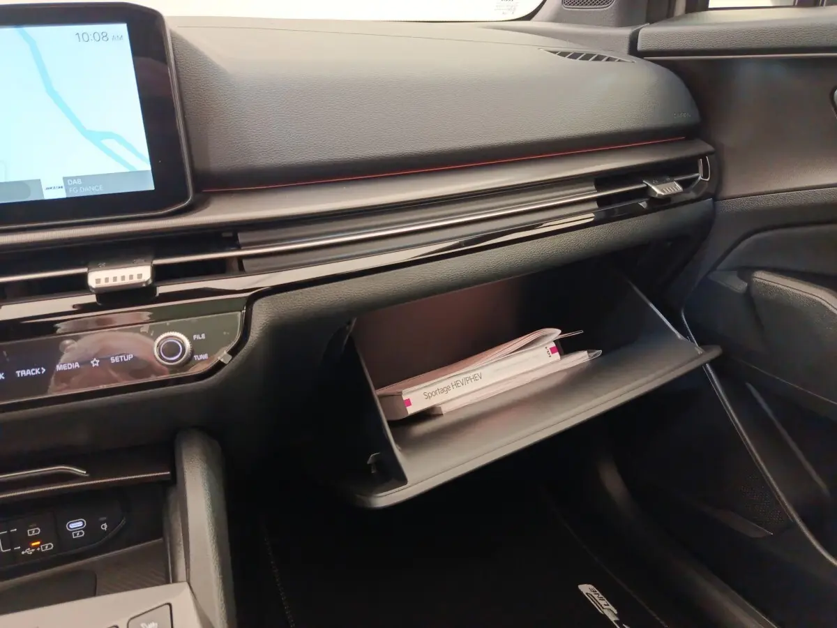Intérieur du Kia Sportage 2026, vue côté passager sur la boîte à gants ouverte avec manuel visible, tableau de bord noir.