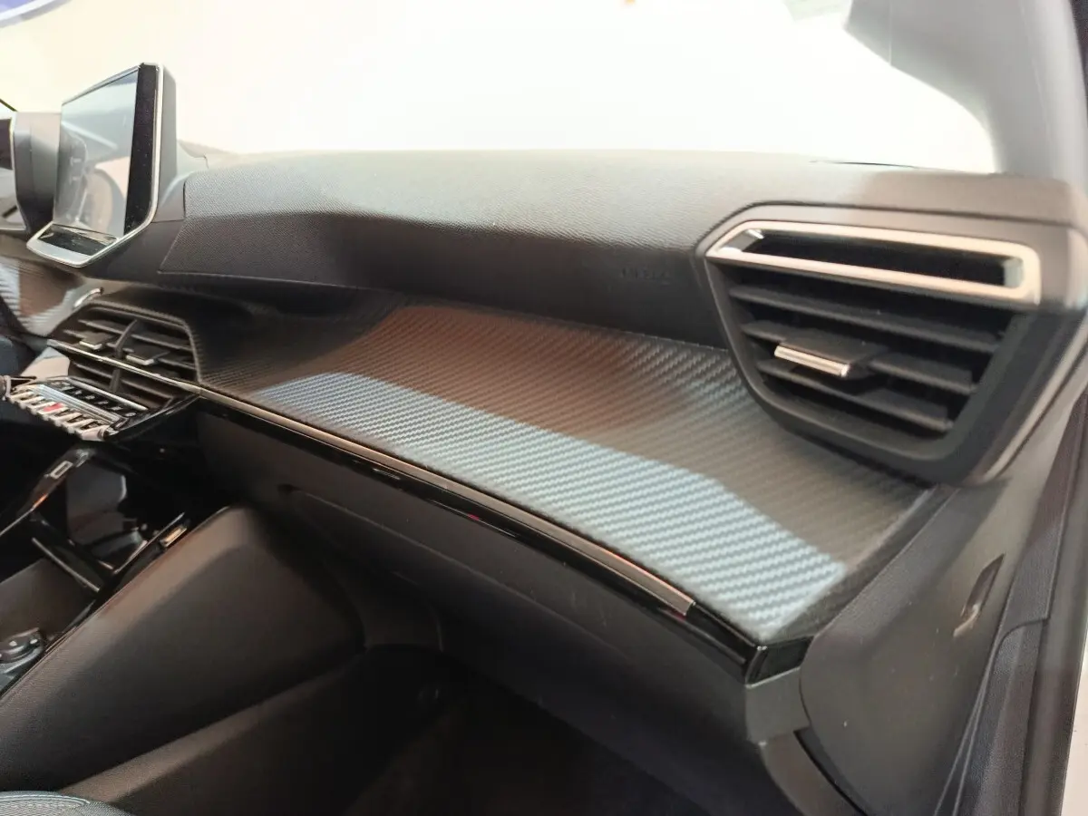 Vue intérieure côté passager du tableau de bord noir avec insert effet carbone du Peugeot 208 PureTech 100 S&S Allure Business.