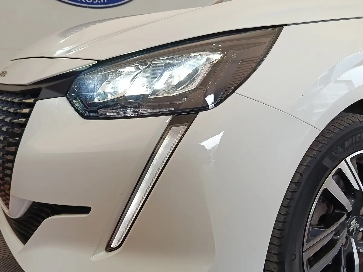 Gros plan sur le phare avant droit allumé d'une Peugeot 208 blanche, avec partie de la calandre et roue visible.