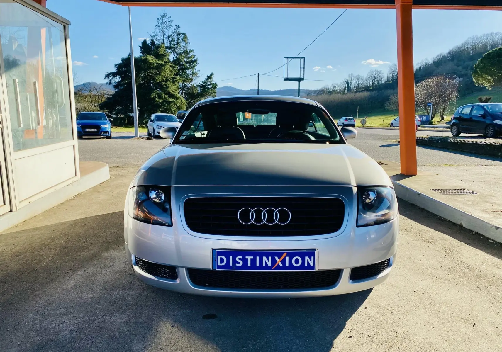Vue frontale d'une Audi TT Coupé gris métallisé garée sous un abri avec plaque Distinxion visible.