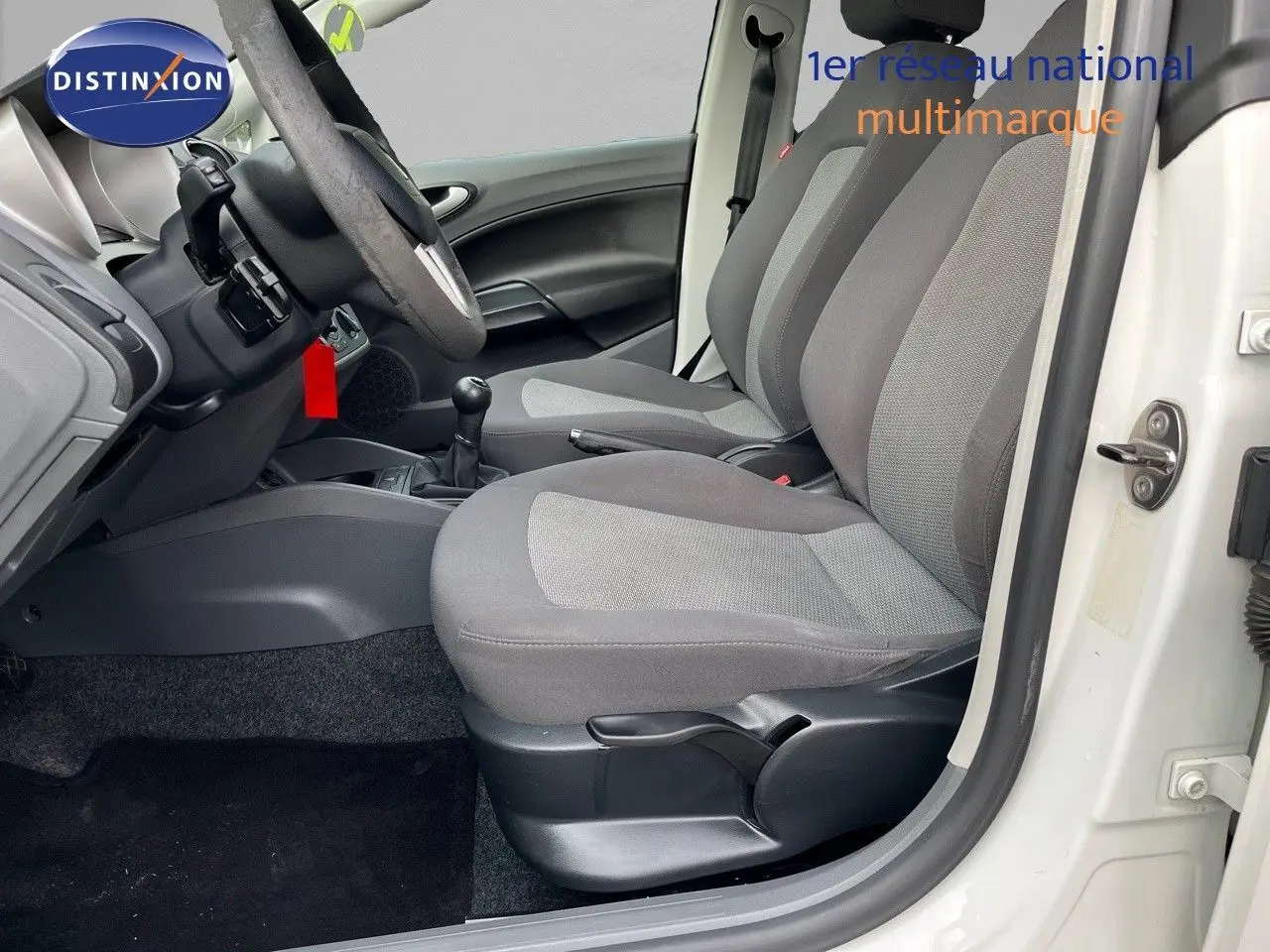 Vue intérieure côté conducteur d'une SEAT Ibiza blanche 2010 avec sièges gris et boîte manuelle visible.