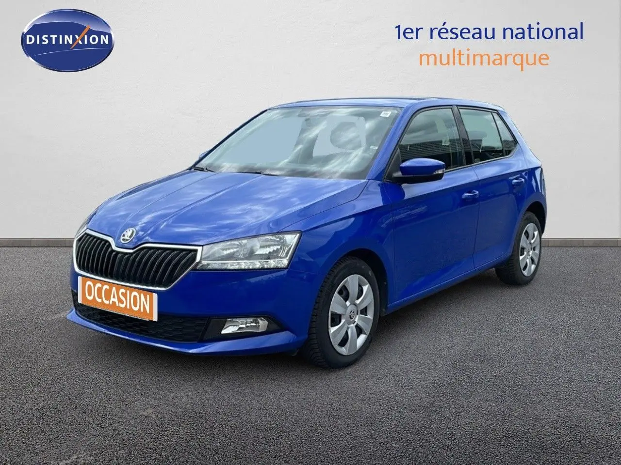 Skoda Fabia 1.0 TSI bleu énergie vue 3/4 avant, garée devant un garage avec façade vitrée et enseignes rouges.