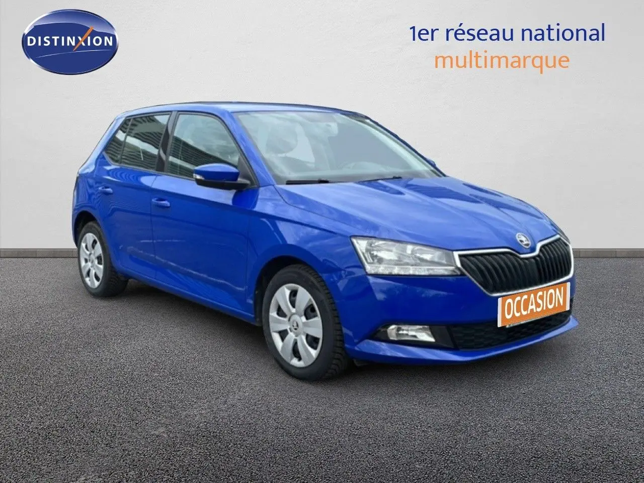Vue avant d'une Skoda Fabia bleu énergie garée devant un garage avec enseigne AD Expert visible.