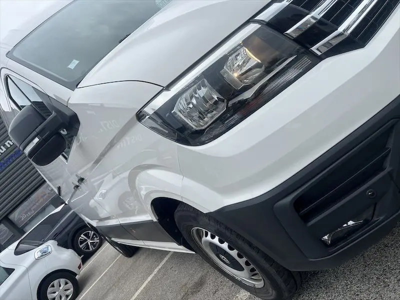 Vue en gros plan 3/4 avant droit du fourgon blanc Volkswagen Crafter L3H3 Business Line 2019 avec phares allumés et rétroviseur noir.