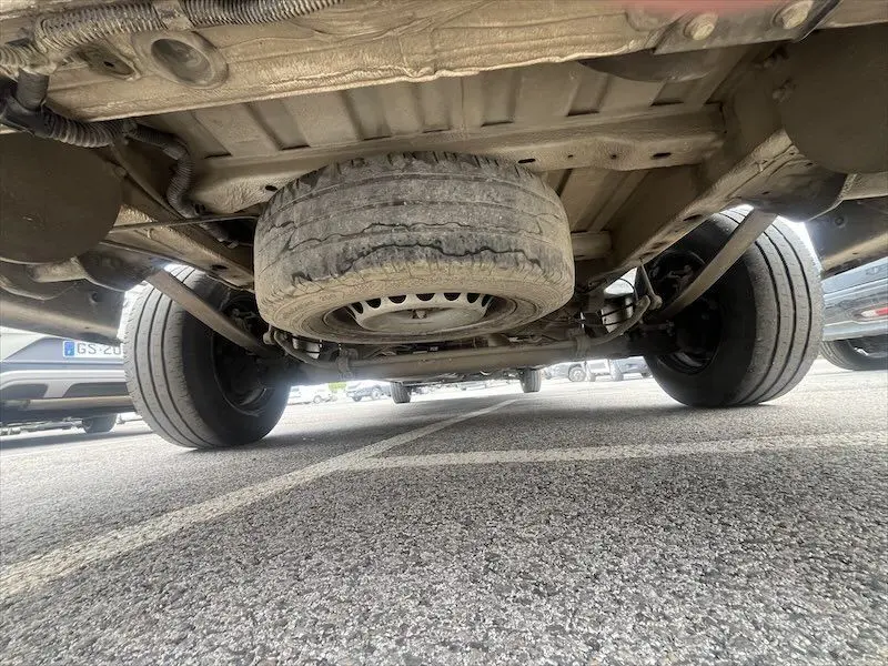 Vue en contre-plongée sous un Volkswagen Crafter blanc montrant la roue de secours et le train arrière sur parking.