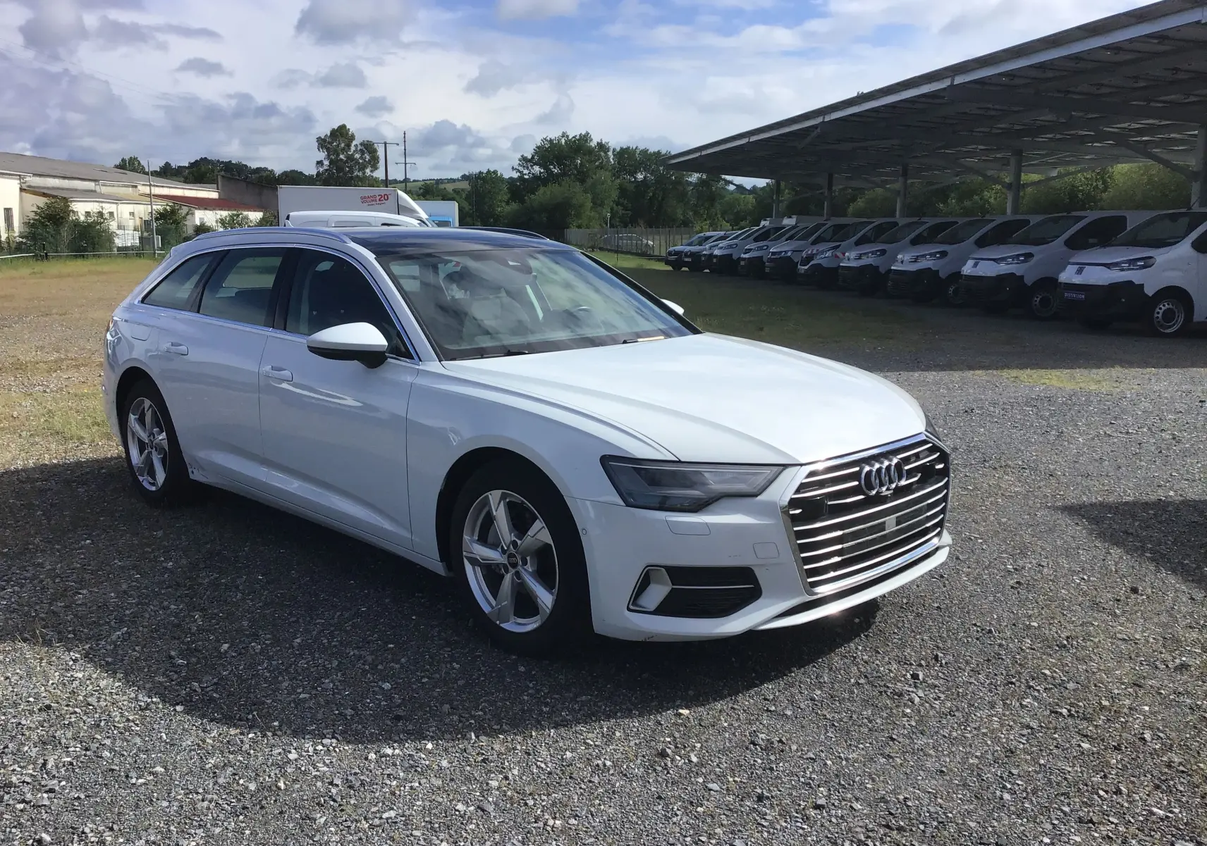 Audi A6 Avant blanc glacier vue 3/4 avant droit sur parking avec rangée de véhicules utilitaires blancs en arrière-plan.