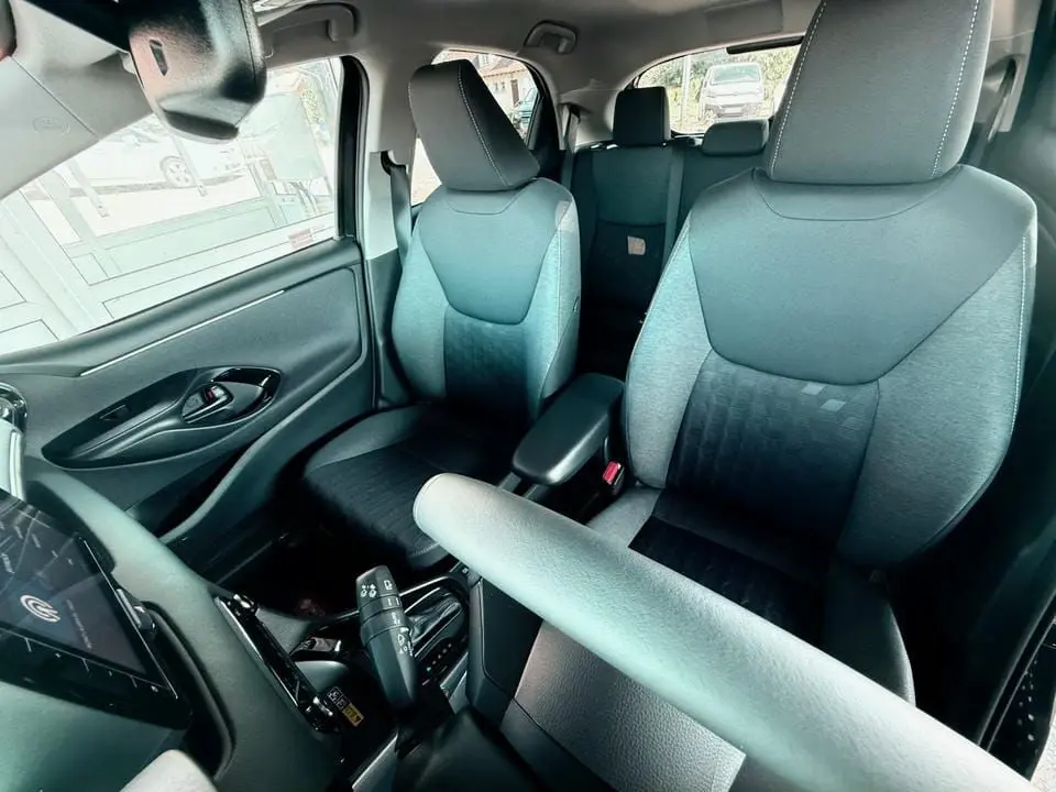 Intérieur de la Toyota Yaris Hybrid 2025 vu côté conducteur, sièges tissu gris et noir avec accoudoir central.