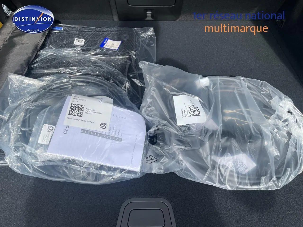 Coffre ouvert d'un Volvo XC60 gris 2024, montrant tapis et accessoires neufs emballés en plastique transparent.