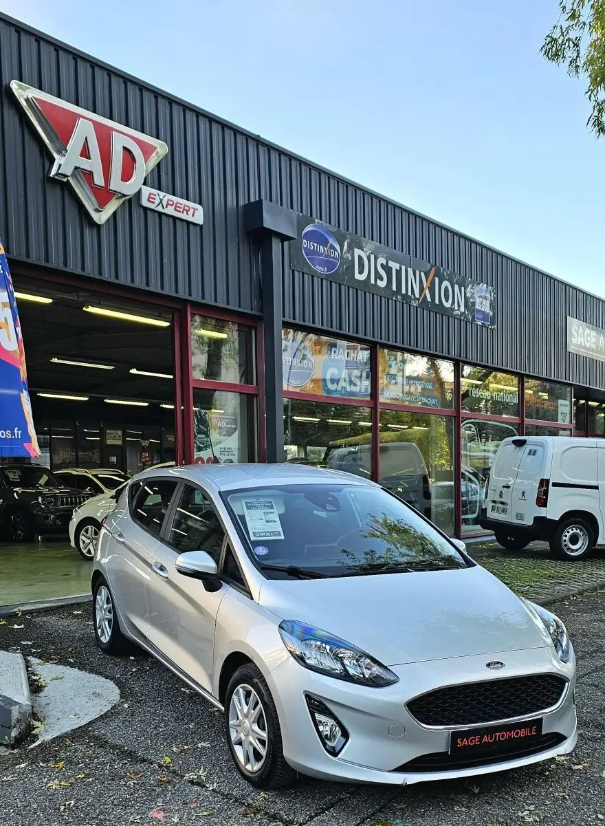 Ford Fiesta gris lunaire métal vue 3/4 avant droit devant un garage avec enseignes commerciales.