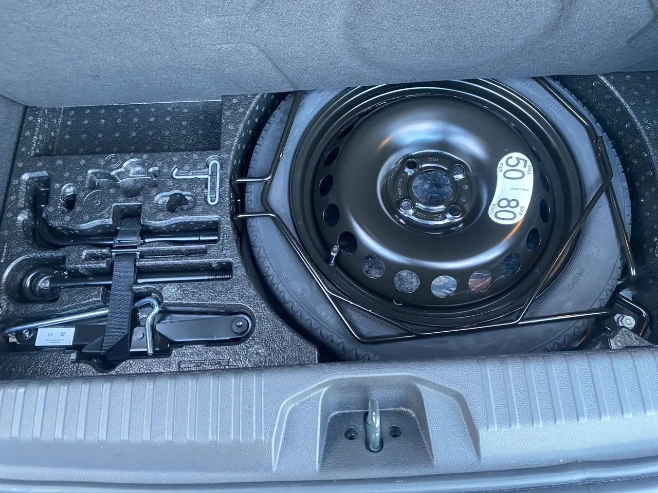 Vue plongeante sur la roue de secours temporaire noire et les outils de bord dans le coffre d'une Renault Clio V gris schiste.