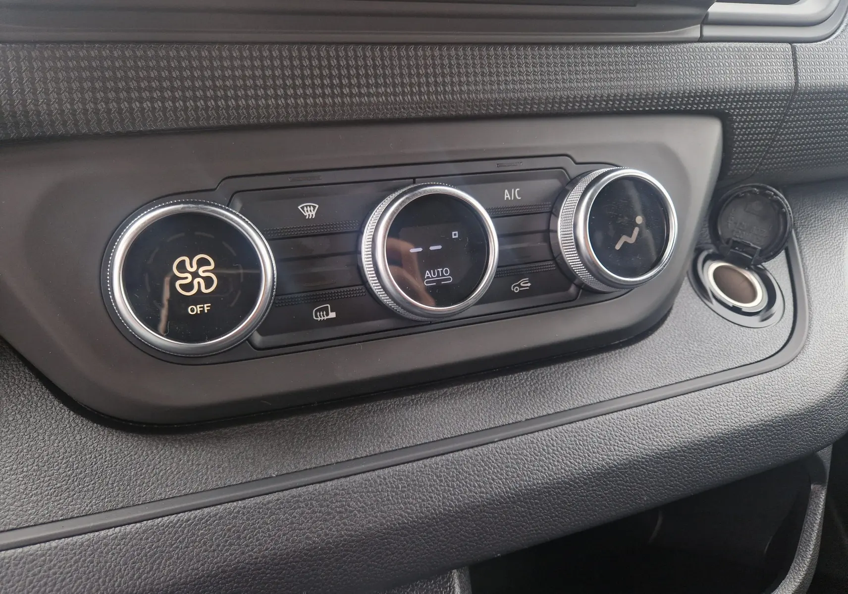 Détail de la console de climatisation du Renault Trafic Fourgon gris urbain, avec commandes rotatives et prise allume-cigare ouverte.