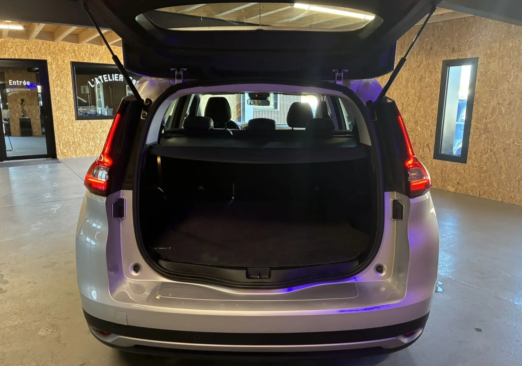 Vue arrière d'un Renault Grand Scenic gris 2017 avec coffre ouvert et feux arrière allumés dans un garage.